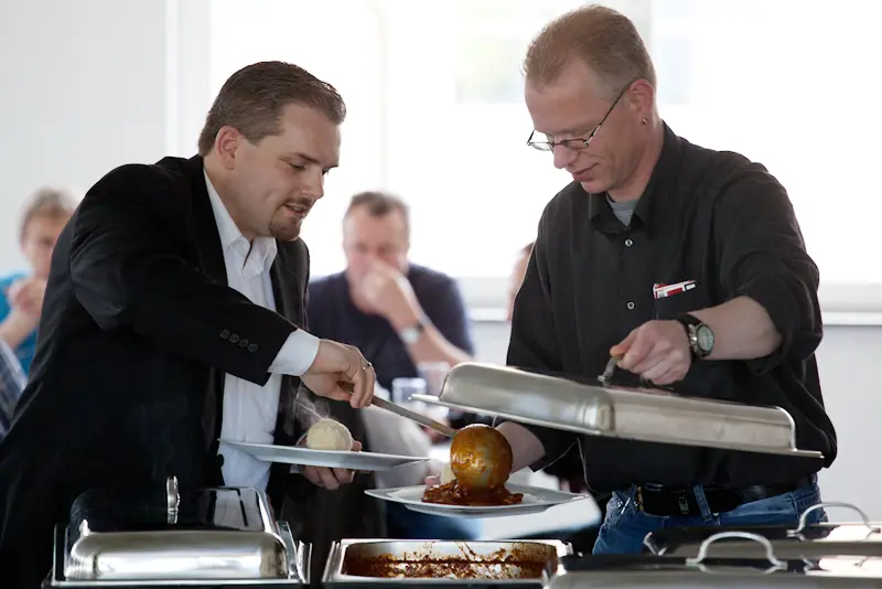 Zwei Männer stehen an einem warmen Buffet, einer hebt den Deckel eines Chafing-Dishes, der andere lässt sich Gulasch mit Kloß auf den Teller geben.