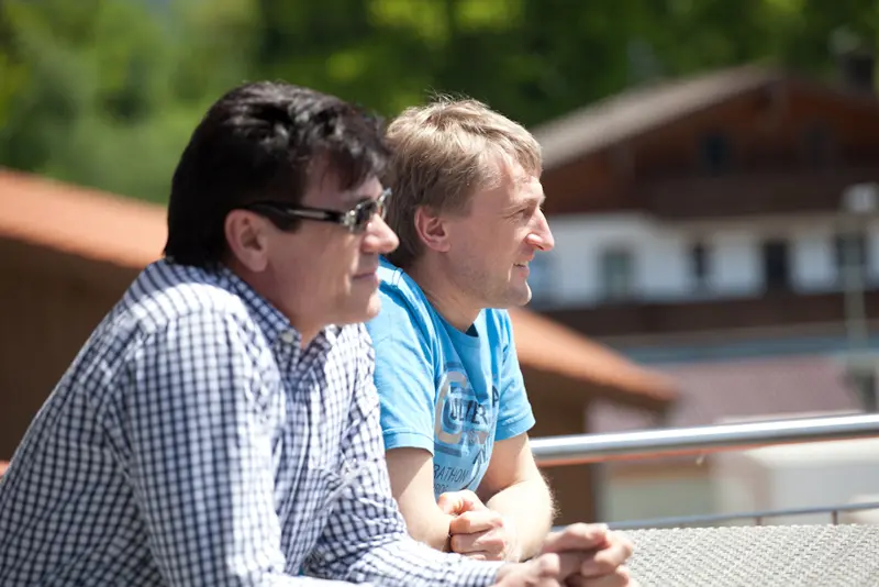 Zwei Männer sitzen entspannt auf einer Terrasse mit Geländer, im Hintergrund sind grüne Bäume und ein Gebäude mit Satteldach zu sehen.
