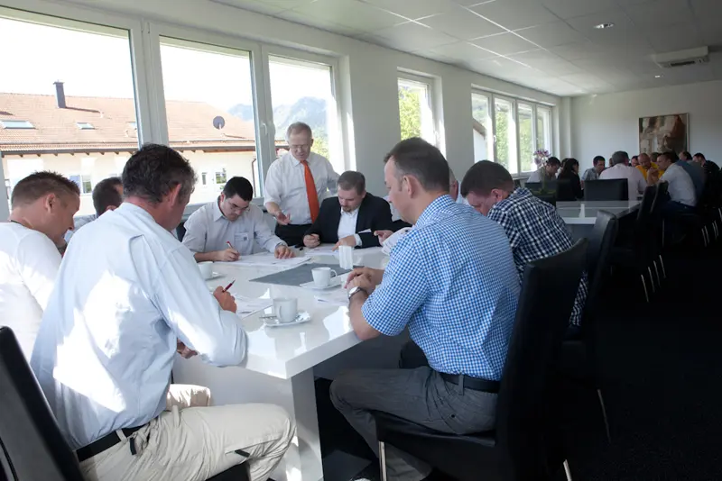 Gruppe von Teilnehmern sitzt mit einem Moderator an einem langen Tisch in einem hellen Raum mit großen Fenstern und arbeitet gemeinsam an Unterlagen.