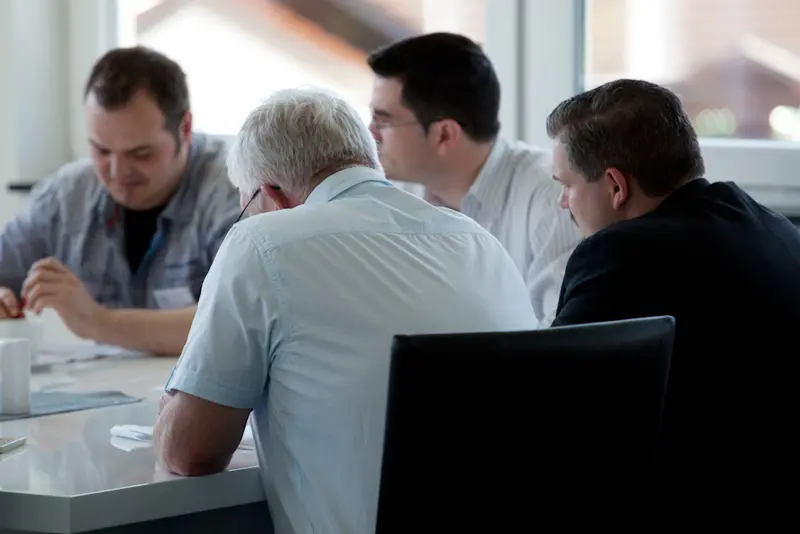 Vier Männer sitzen in einer Kleingruppe an einem Tisch und arbeiten konzentriert an Unterlagen in einem hellen Raum mit großen Fenstern.