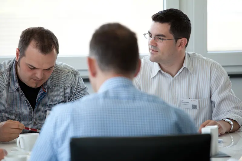 Drei Seminarteilnehmer sitzen an einem Tisch, zwei arbeiten an Unterlagen, einer hört aufmerksam zu, im Hintergrund helle Fensterfront.