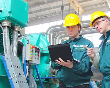 Two maintenance technicians wearing helmets check data on a laptop in front of a large industrial plant as part of lubricant management.