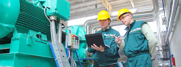 Two maintenance technicians wearing helmets check data on a laptop in front of a large industrial plant as part of lubricant management.